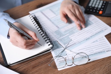 Payroll. Woman taking notes in notebook while working with tax return forms at wooden table, closeup