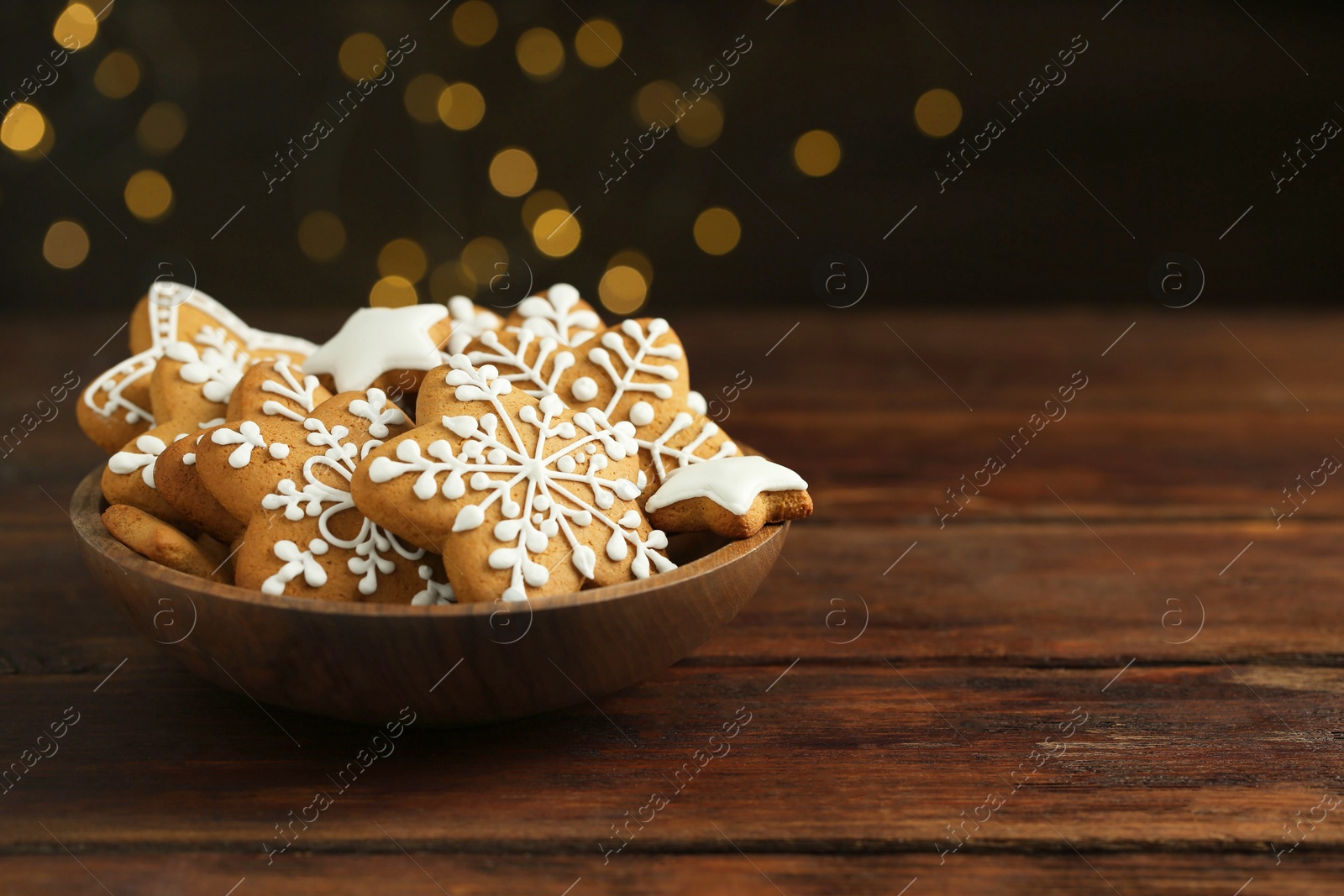 Photo of Tasty Christmas cookies with icing in bowl on wooden table against blurred lights. Space for text