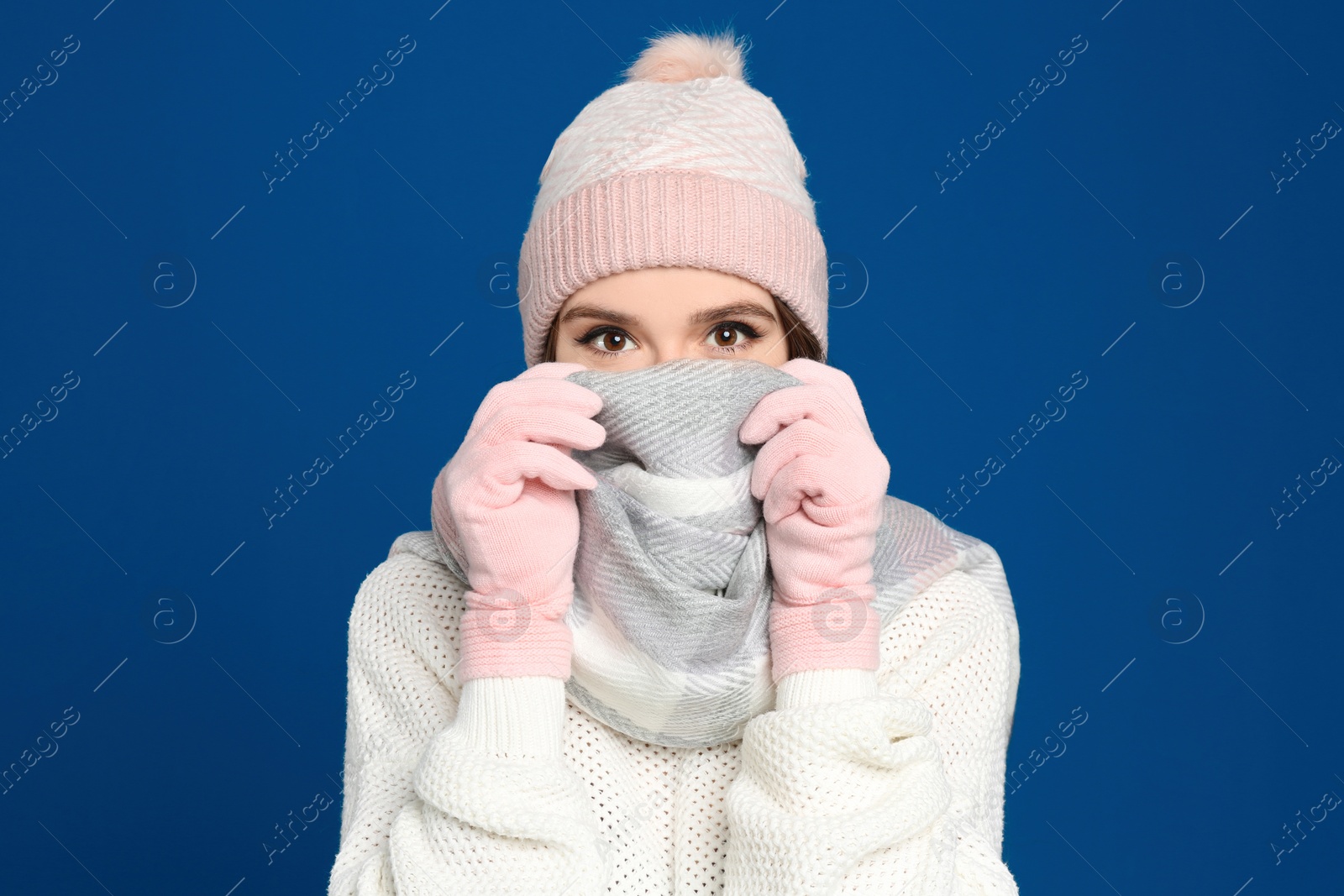 Photo of Young woman wearing warm sweater, gloves, scarf and hat on blue background. Winter season