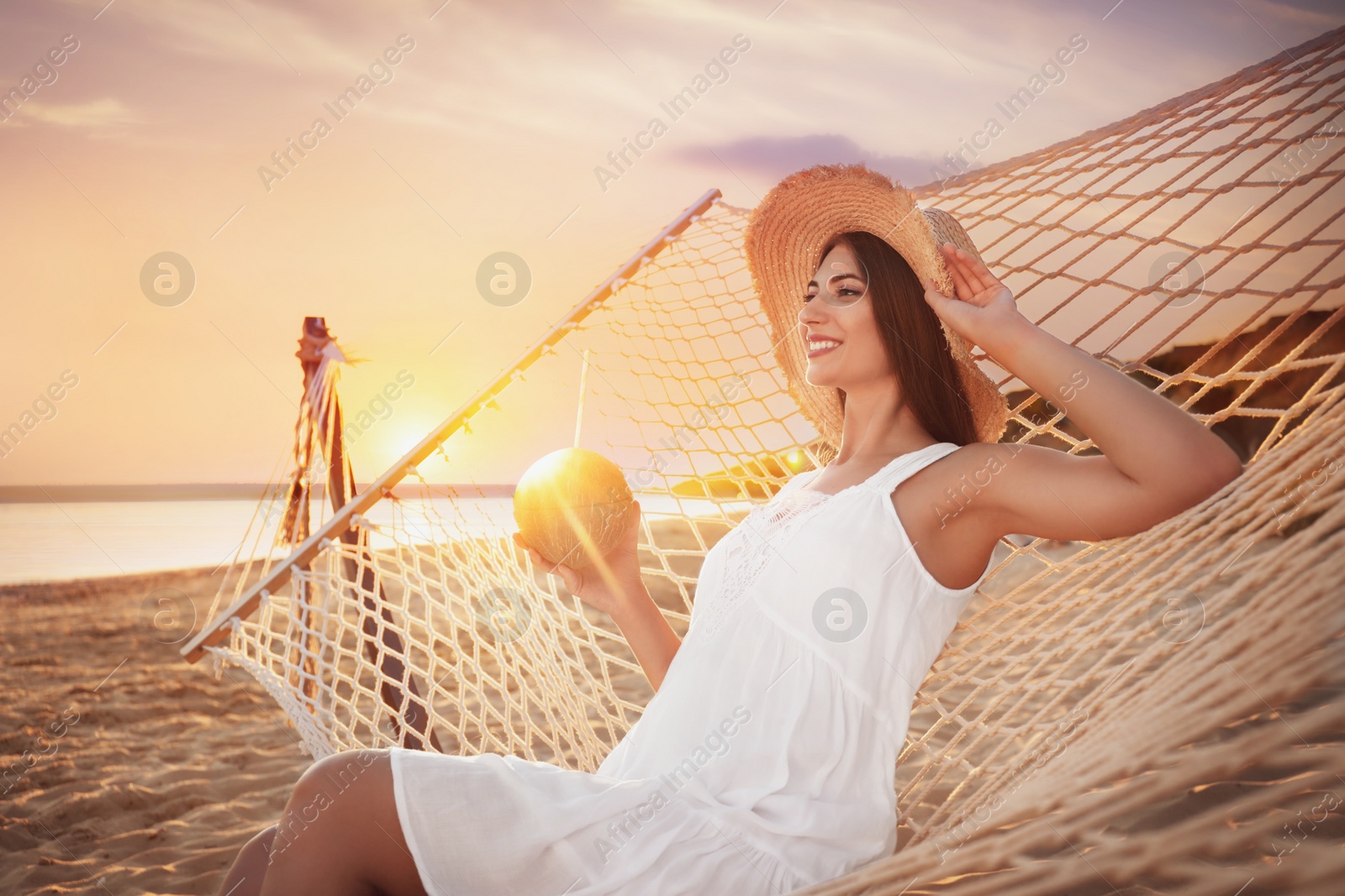 Photo of Young woman with tropical cocktail relaxing in hammock on beach at sunset