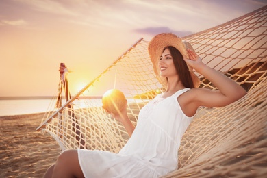 Young woman with tropical cocktail relaxing in hammock on beach at sunset