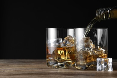 Photo of Pouring whiskey into glass with ice cubes at wooden table against black background, space for text