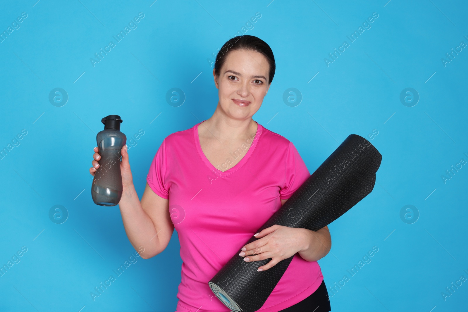 Photo of Happy overweight woman with bottle of water and yoga mat on light blue background