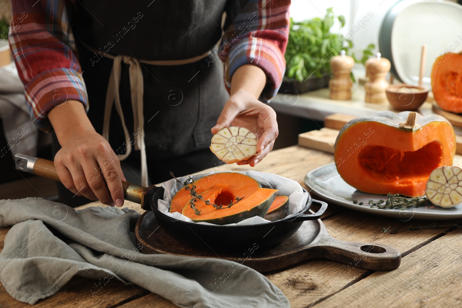 Photo of Woman adding thyme and garlic to pumpkin slices in frying pan at wooden table in kitchen, closeup