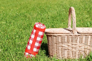 Rolled checkered tablecloth near picnic basket on green grass outdoors, space for text