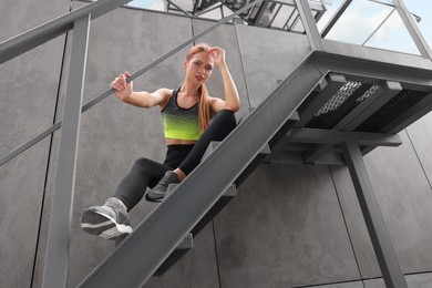 Photo of Beautiful woman in stylish gym clothes sitting on stairs on street, low angle view