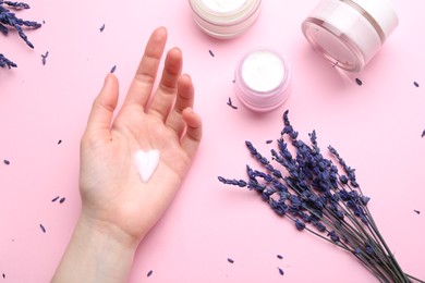 Woman applying hand cream and lavender flowers on pink background, top view