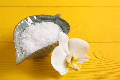 Photo of Natural sea salt in bowl and orchid flower on yellow wooden table. Space for text