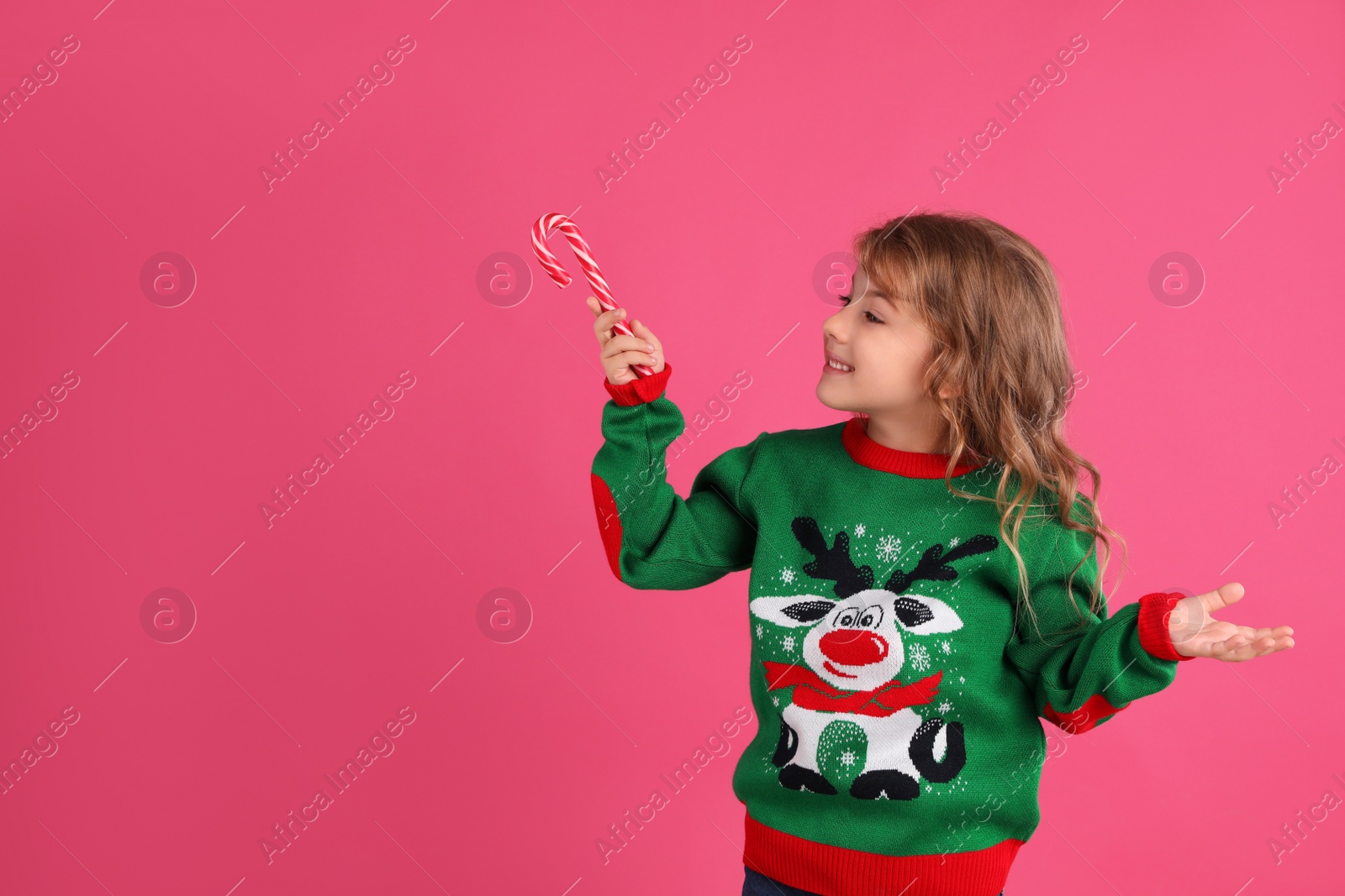 Photo of Cute little girl in green Christmas sweater holding candy cane against pink background. Space for text