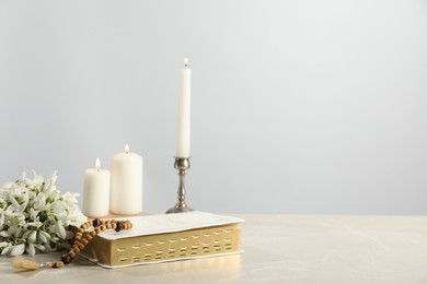 Photo of Church candles, wooden cross, rosary beads, Bible and flowers on light table. Space for text