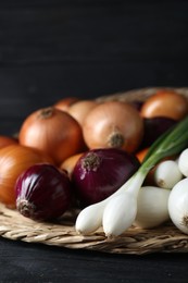 Photo of Wicker mat with different kinds of onions on black wooden table, closeup