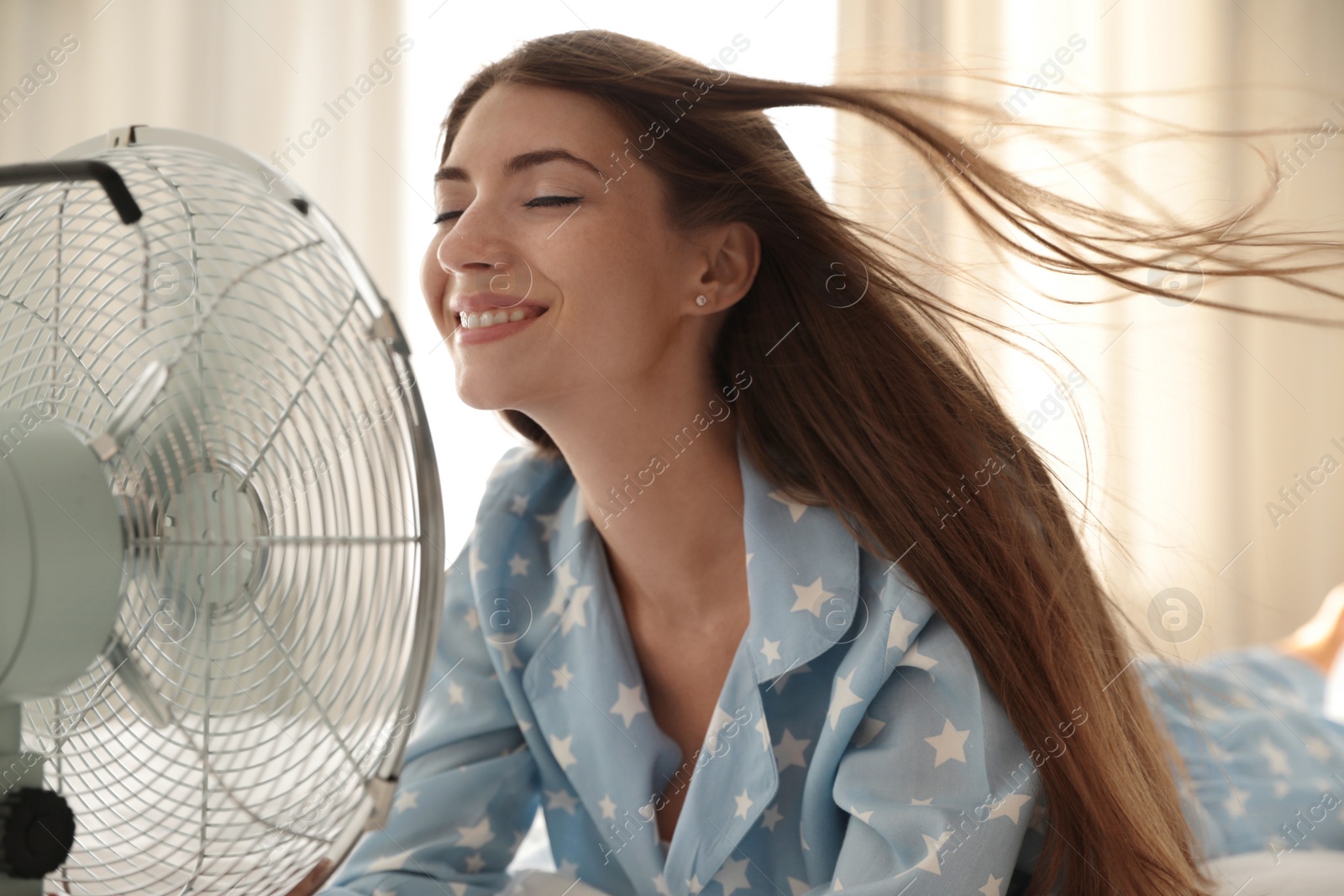 Photo of Woman enjoying air flow from fan on bed in room. Summer heat