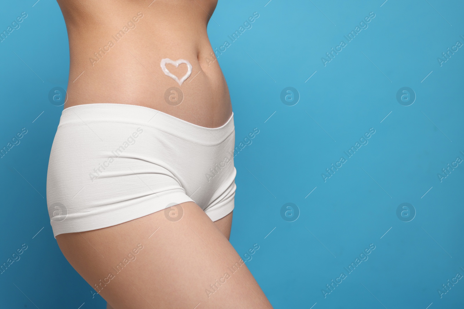 Photo of Woman with heart made of body cream on her belly against light blue background, closeup. Space for text