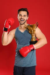 Photo of Portrait of happy young boxer with gold trophy cup on red background