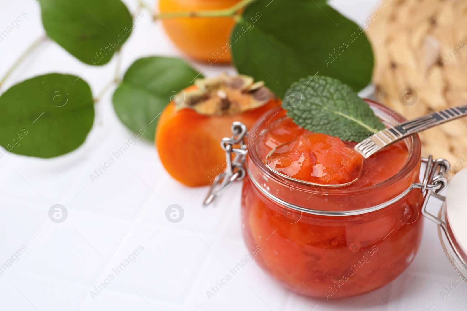 Photo of Jar and spoon of tasty persimmon jam, ingredients on white tiled table. Space for text