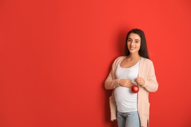 Happy pregnant woman with Christmas ball on red background, space for text. Expecting baby