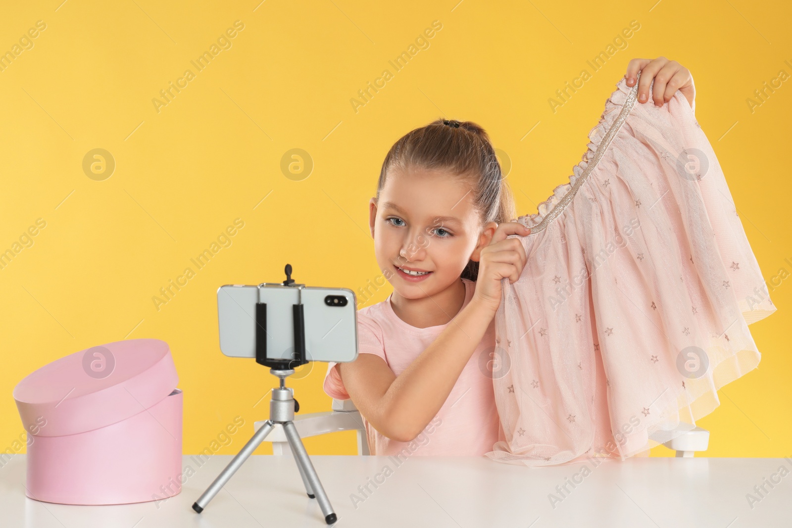 Photo of Cute little blogger with skirt recording video at table on yellow background