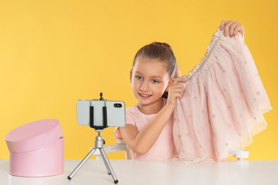 Photo of Cute little blogger with skirt recording video at table on yellow background