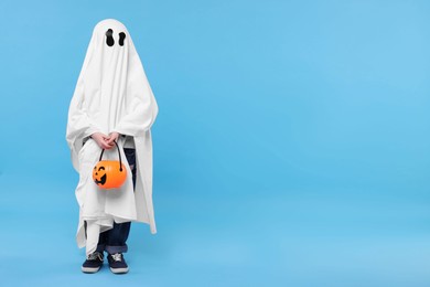 Photo of Child in white ghost costume holding pumpkin bucket on light blue background, space for text. Halloween celebration