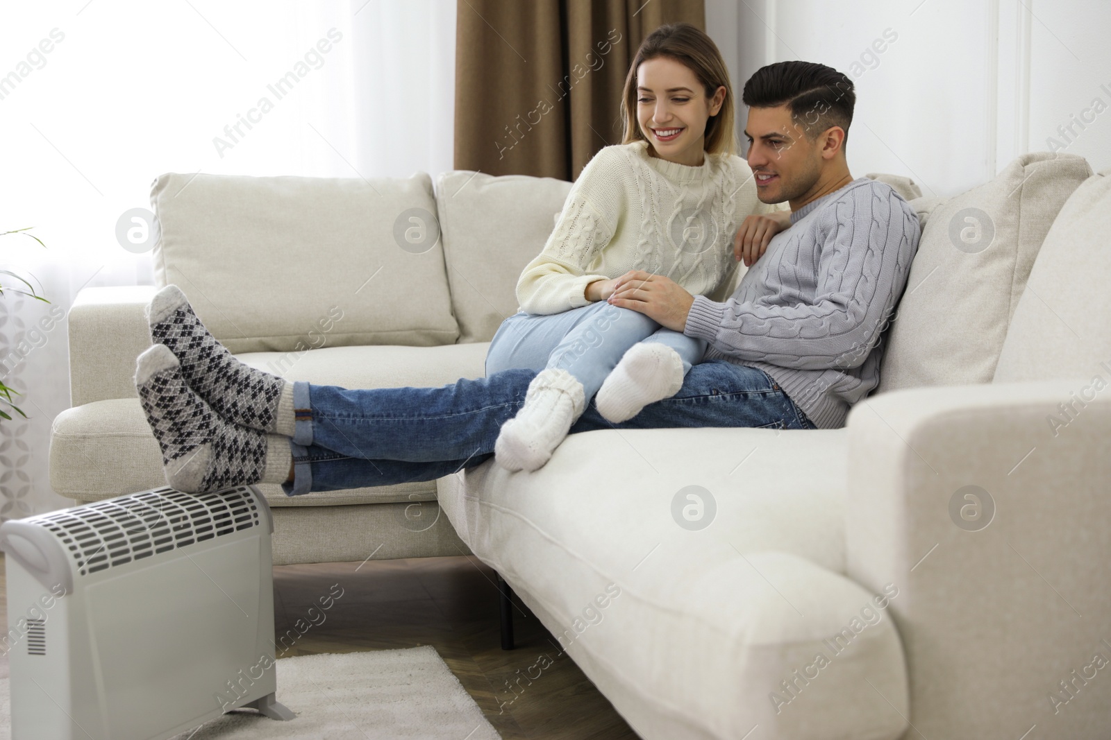 Photo of Happy couple sitting on sofa near electric heater at home