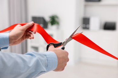 Photo of Man cutting red ribbon with scissors indoors, closeup
