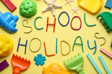 Photo of Text School Holidays made of modelling clay and different sand toys on yellow background, flat lay