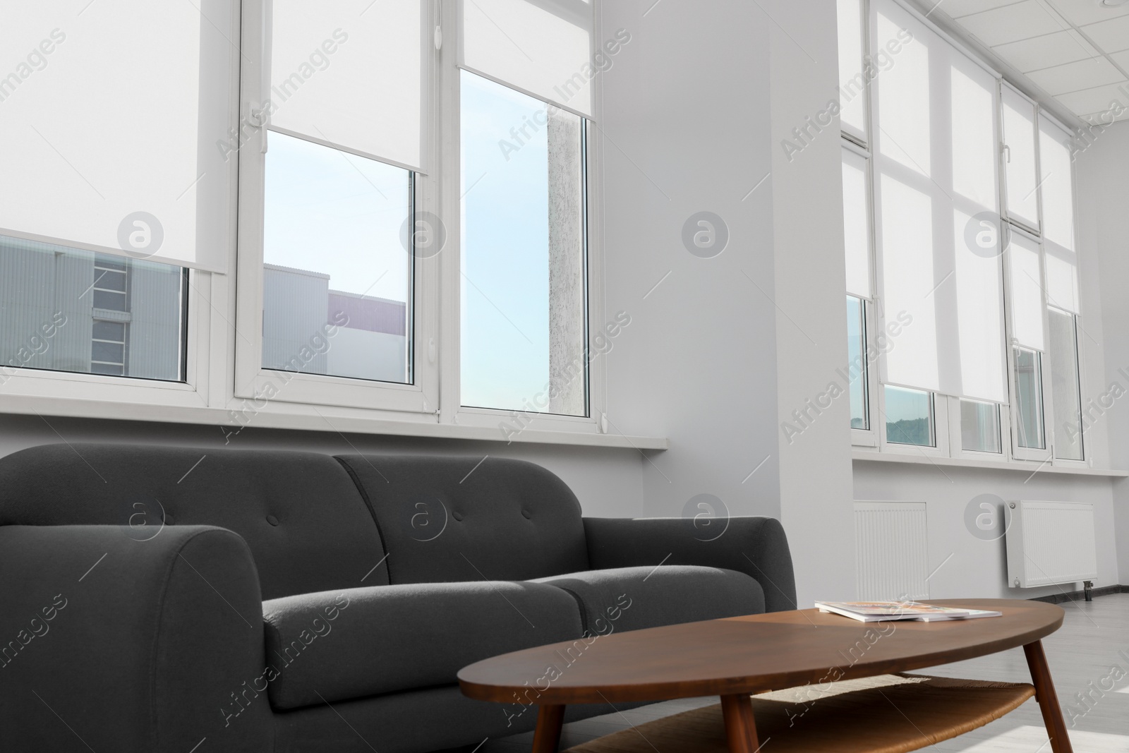 Photo of Comfortable sofa and table near large windows with white roller blinds indoors