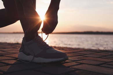 Photo of Young woman tying shoelaces near river at sunrise, space for text. Morning training