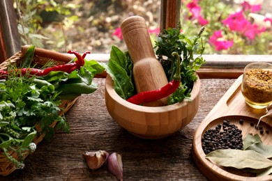 Photo of Mortar with pestle, fresh green herbs and different spices on wooden table near window