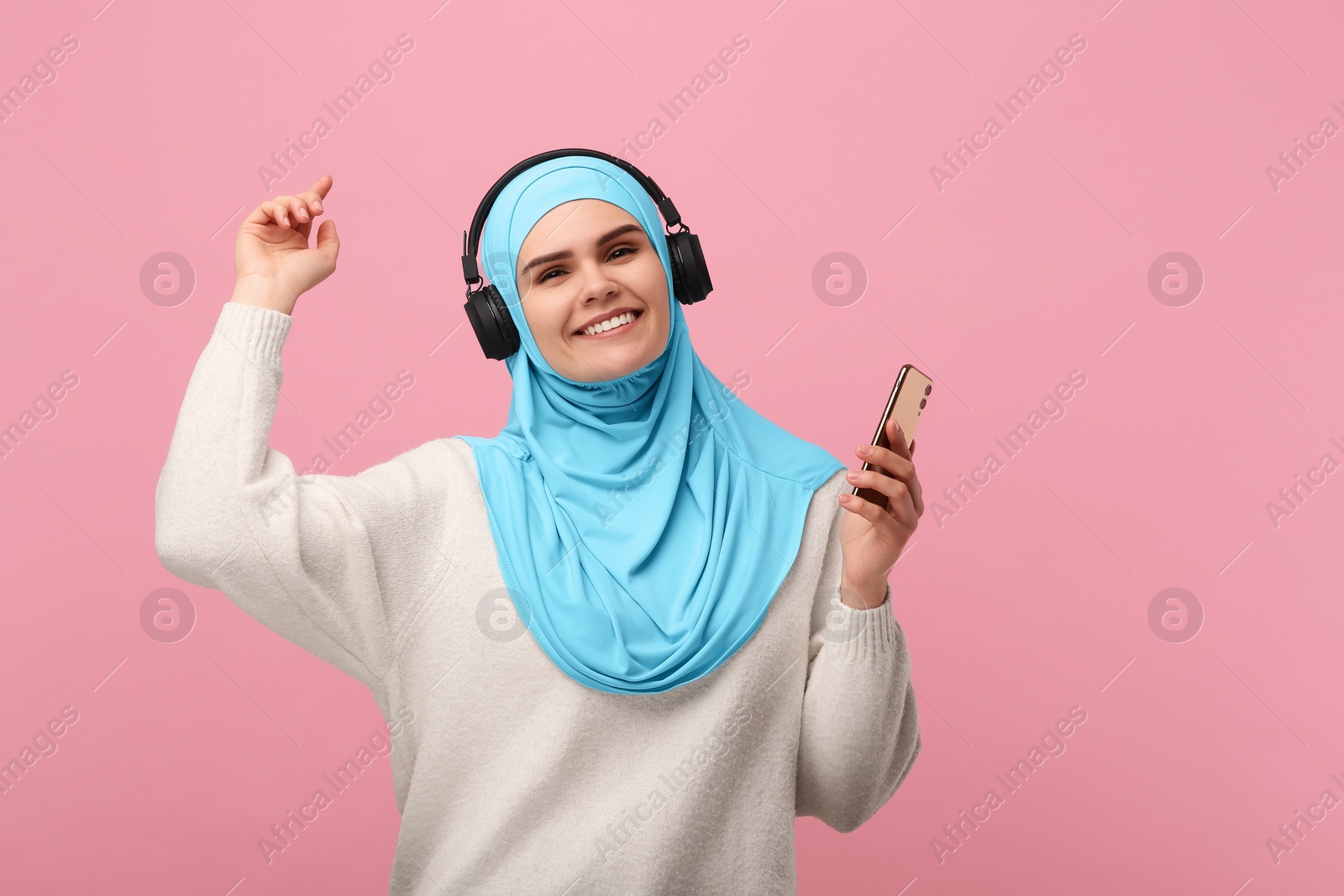 Photo of Muslim woman in hijab and headphones dancing with smartphone on pink background