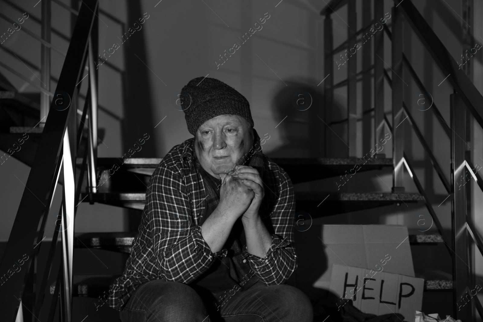 Photo of Poor senior man with cardboard sign HELP on stairs indoors. Black and white effect