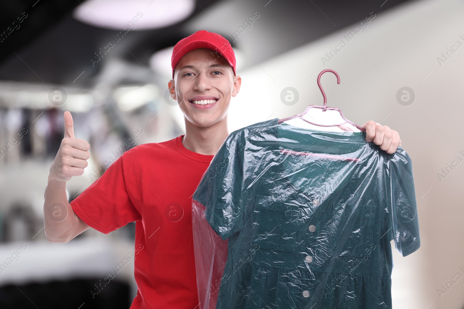 Image of Happy courier holding dress in plastic bag and showing thumbs up in dry-cleaning
