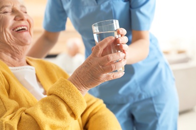 Photo of Nurse giving glass of water to senior woman indoors, closeup. Assisting elderly people