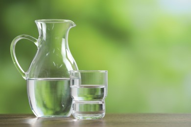 Photo of Jug and glass with clear water on wooden table against blurred green background, closeup. Space for text