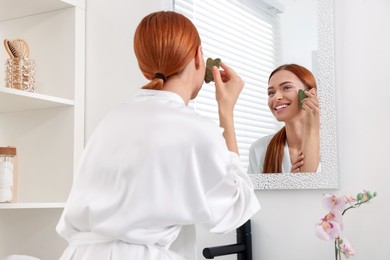 Photo of Young woman massaging her face with jade gua sha tool near mirror in bathroom