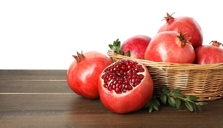 Photo of Fresh pomegranates in wicker basket and green leaves on wooden table against white background, space for text