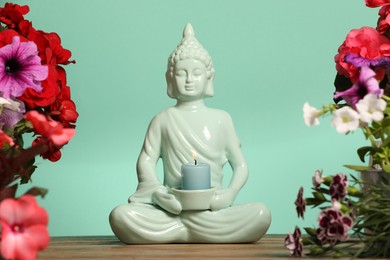 Photo of Buddhism religion. Decorative Buddha statue with burning candle and beautiful flowers on table against turquoise wall, selective focus