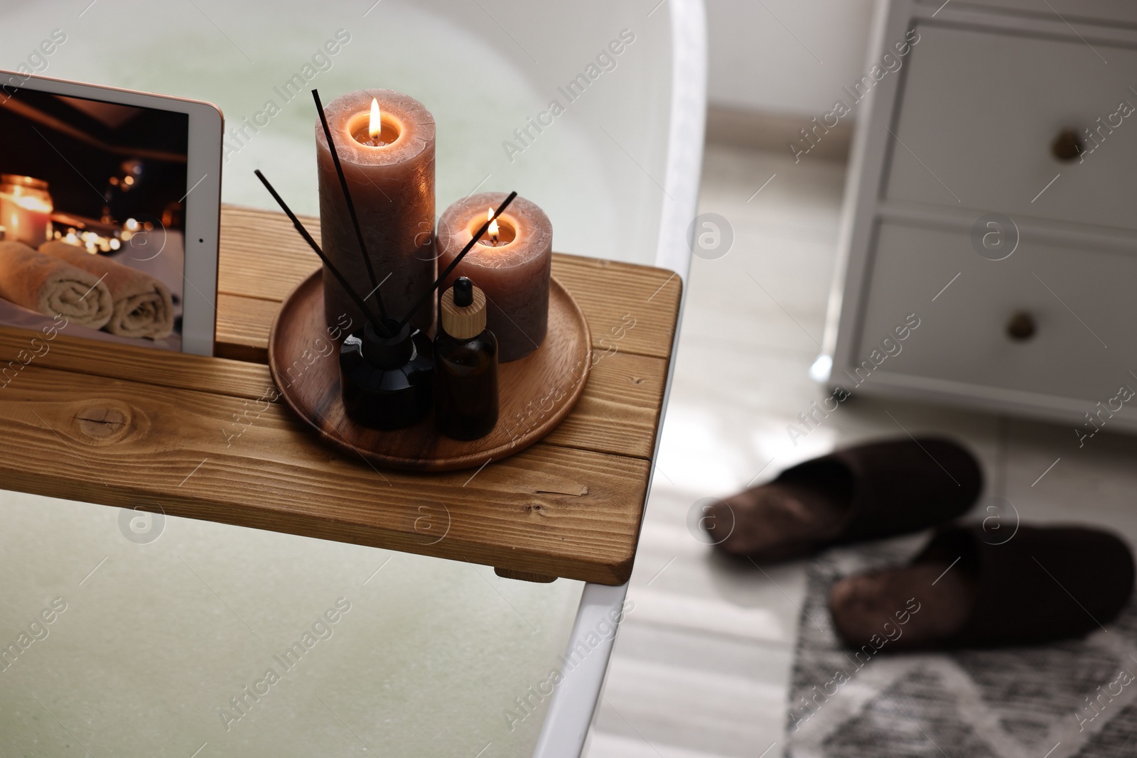 Photo of Wooden tray with tablet, burning candles and aroma products on bathtub in bathroom. Space for text