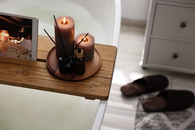 Photo of Wooden tray with tablet, burning candles and aroma products on bathtub in bathroom. Space for text