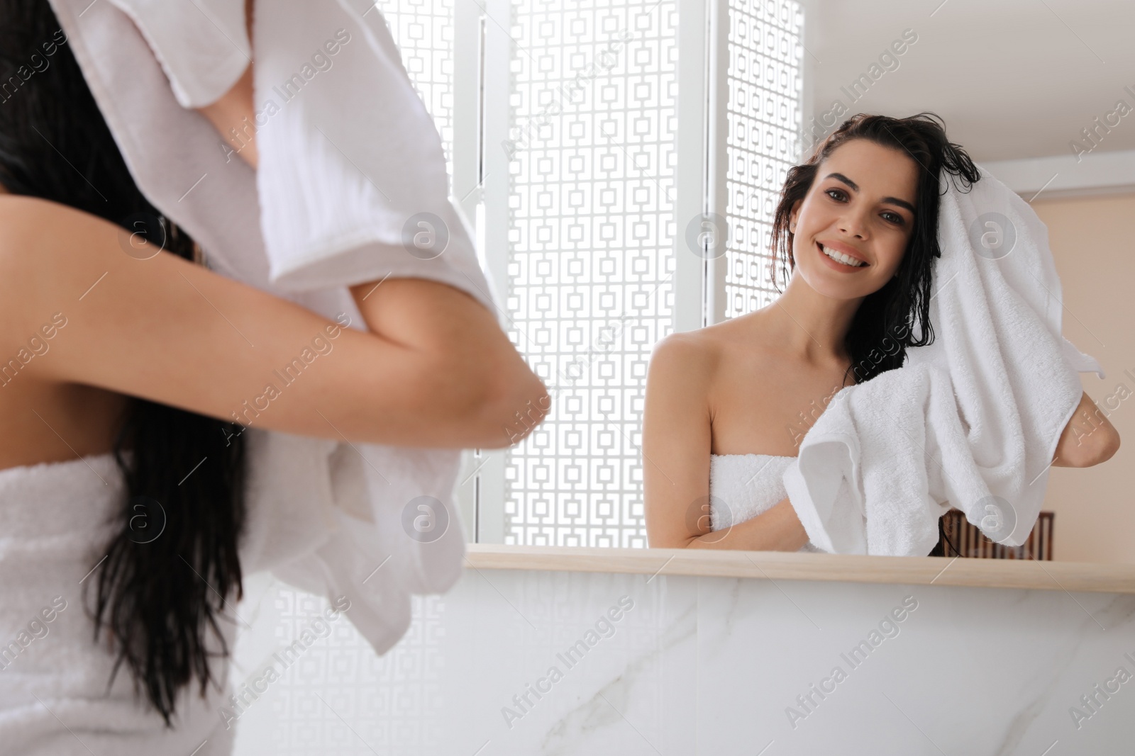 Photo of Happy young woman drying hair with towel after washing near mirror in bathroom