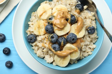 Tasty oatmeal with banana, blueberries and peanut butter served in bowl on light blue wooden table, flat lay