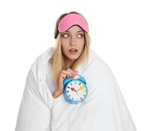 Emotional woman in sleeping mask wrapped with blanket holding alarm clock on white background
