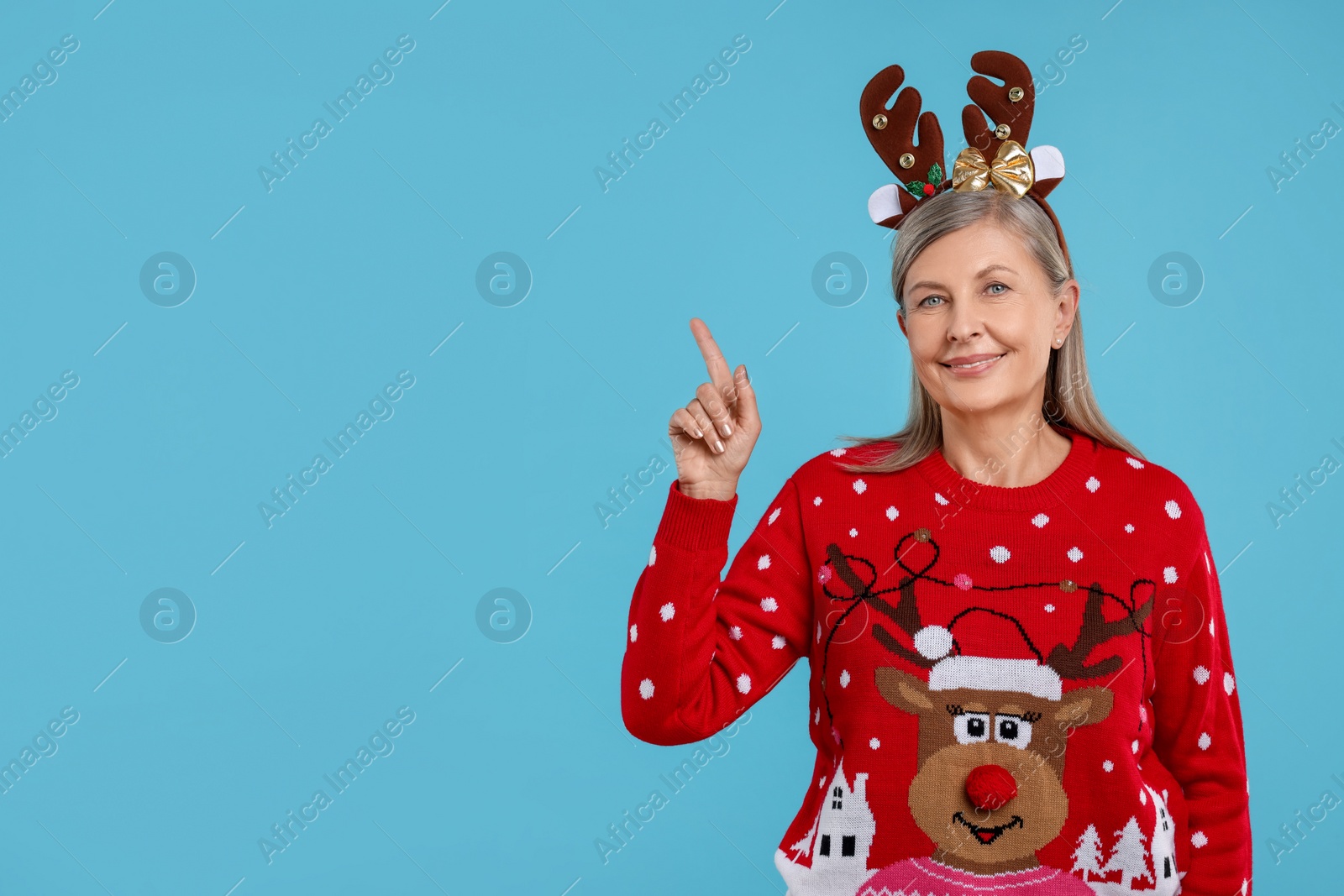 Photo of Senior woman in Christmas sweater and reindeer headband pointing at something on light blue background. Space for text