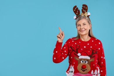 Photo of Senior woman in Christmas sweater and reindeer headband pointing at something on light blue background. Space for text