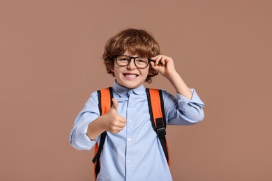 Photo of Happy schoolboy in glasses with backpack showing thumb up gesture on brown background