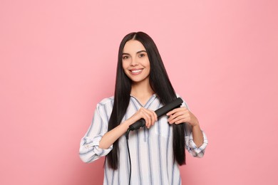 Beautiful happy woman using hair iron on pink background