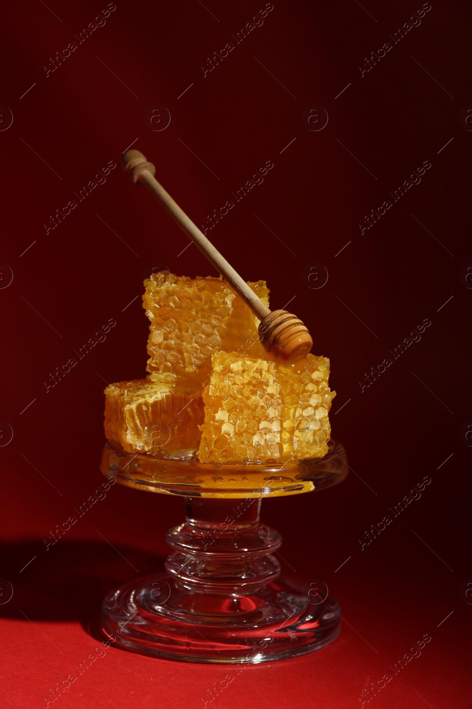 Photo of Natural honeycombs and wooden dipper on glass stand against burgundy background