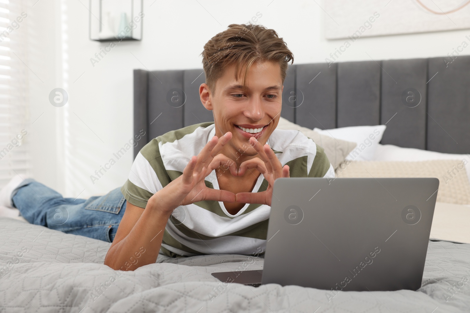 Photo of Happy young man having video chat via laptop and making heart with hands on bed indoors. Long-distance relationship