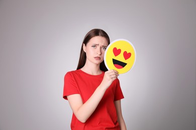 Photo of Sad young woman covering face with heart eyes emoji on grey background