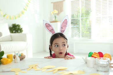 Emotional little girl wearing bunny ears headband at table with Easter eggs, indoors